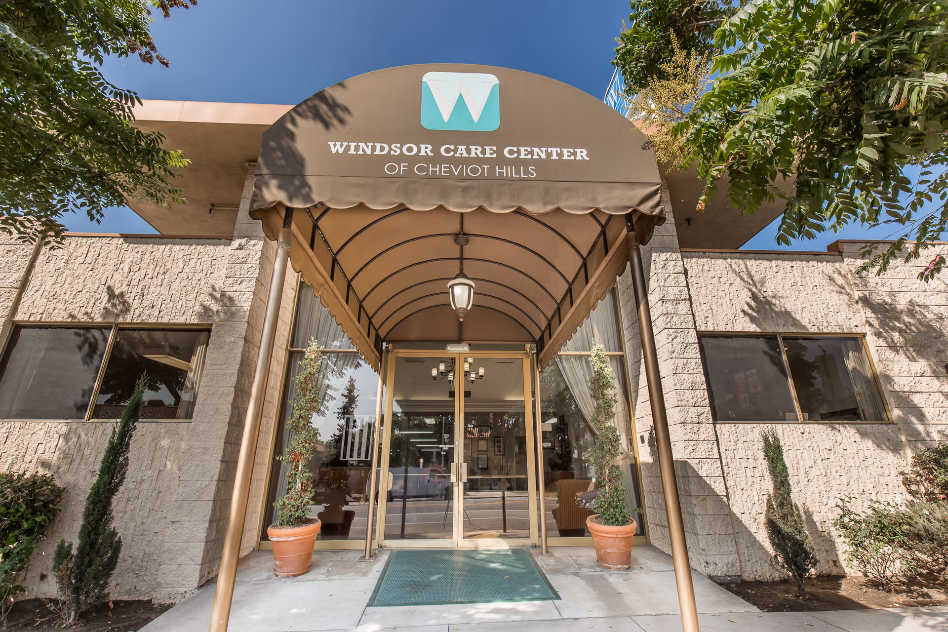 Entrance to Windsor Care Center of Cheviot Hills with a brown canopy displaying the facility name and logo above glass double doors, flanked by potted plants and windows on a sunny day.