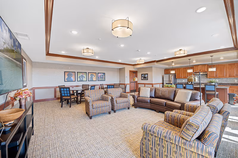 Spacious senior living common room with sofas, armchairs, dining tables, and a kitchenette area.