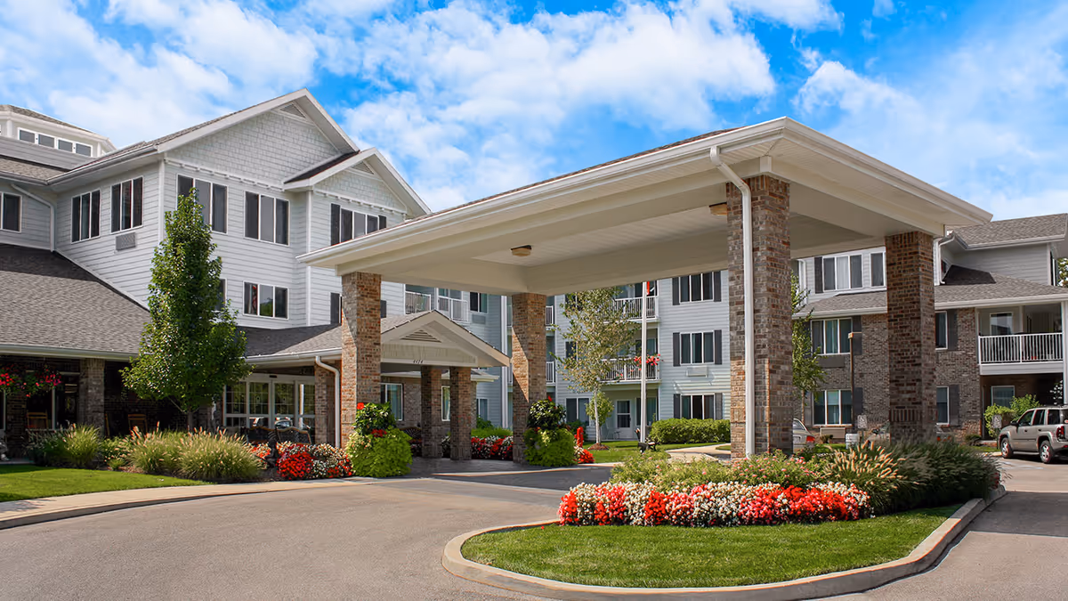 Front entrance and porte-cochère of a multi-story senior living building with landscaped flowerbeds and a driveway.