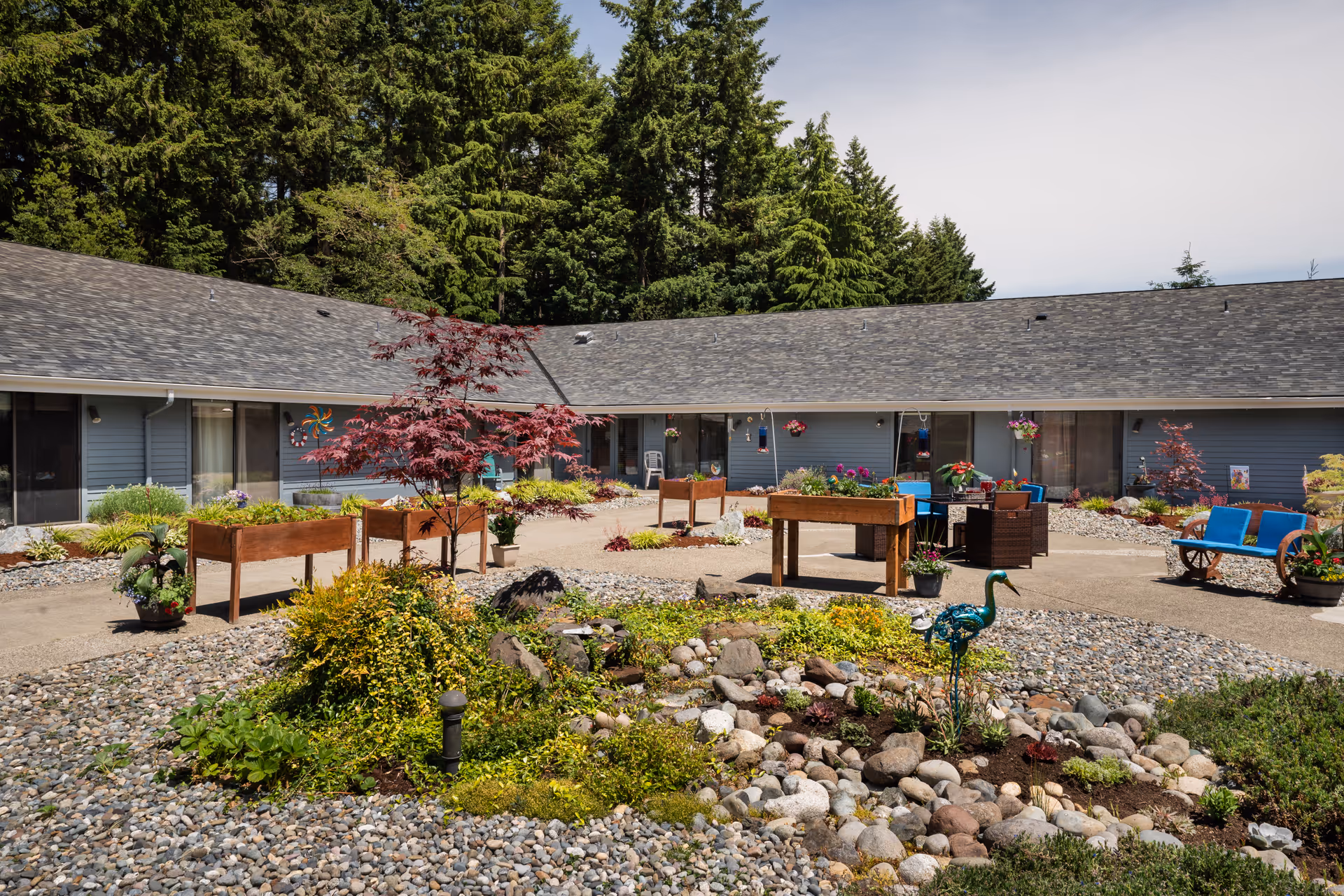 Central courtyard with raised garden beds, seating areas, decorative plants and a single-story building backed by trees.