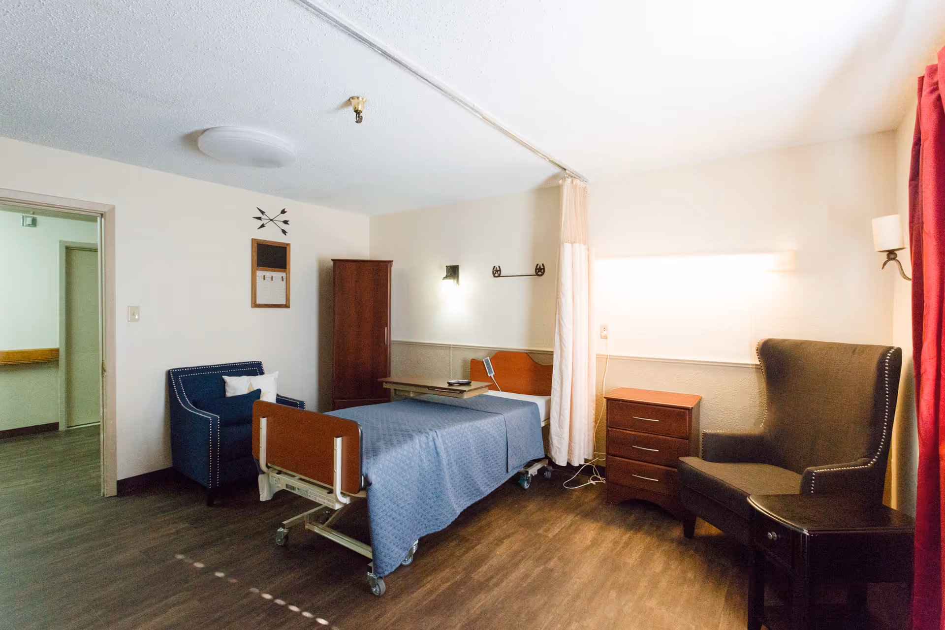 A senior living facility room with a hospital-style bed covered in a blue blanket, a wooden nightstand with three drawers, a blue armchair, a dark wingback chair, a small side table, a wooden wardrobe, and a privacy curtain. The room has wood flooring and light-colored walls with a ceiling light fixture.