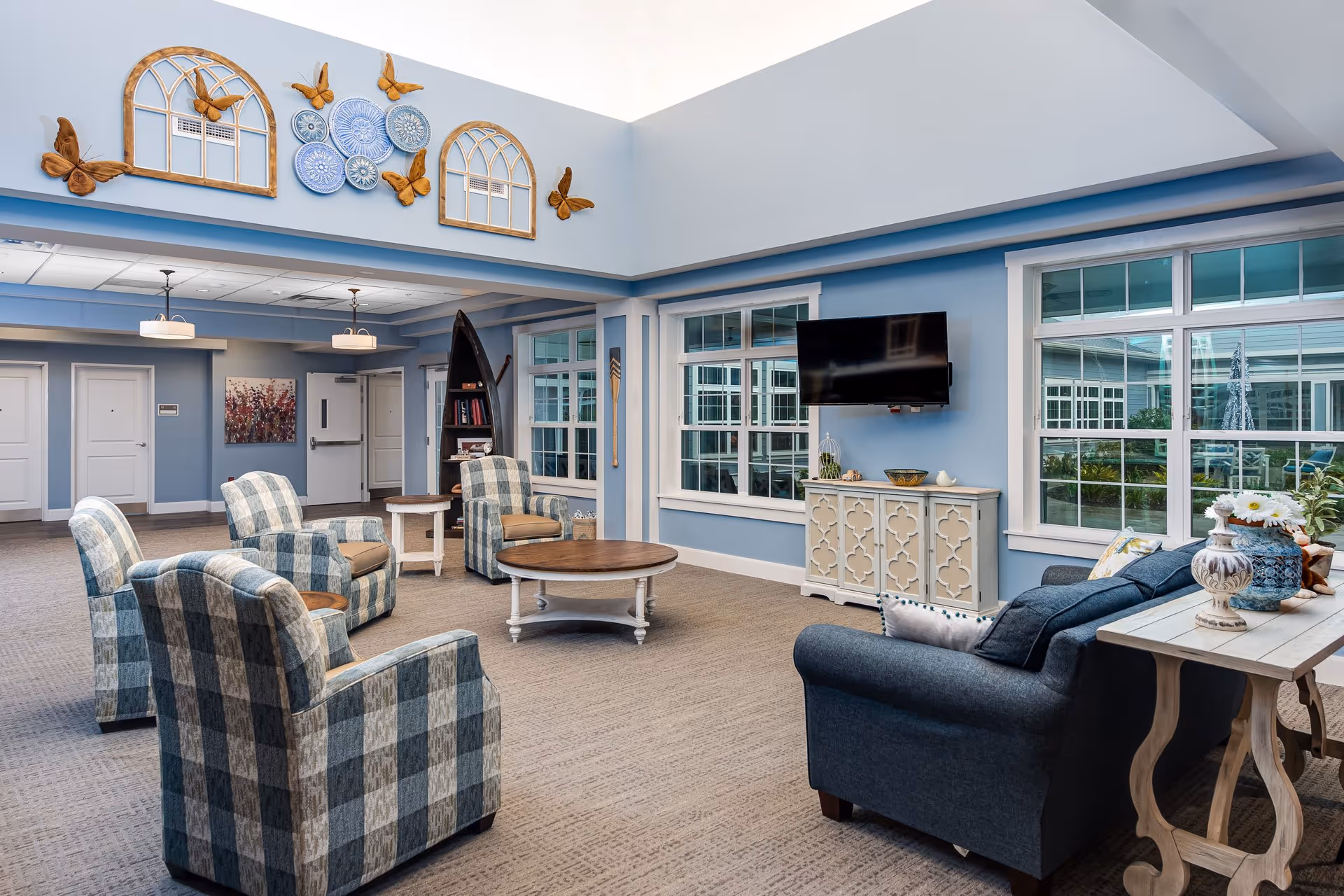 Bright, blue-themed common room with upholstered armchairs and a sofa arranged around coffee tables, a wall-mounted TV, and large windows.