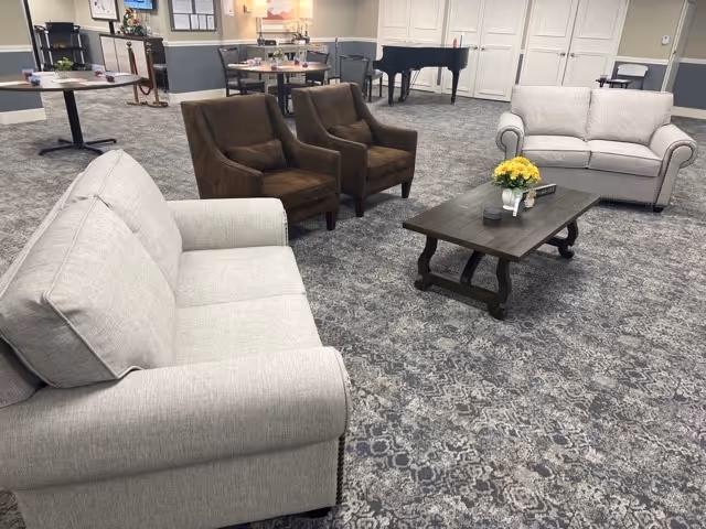 A senior living facility common area with two light gray sofas, two brown armchairs, and a dark wooden coffee table with a flower arrangement and small decorative items. In the background, there are tables and chairs, a piano, and white paneled doors.
