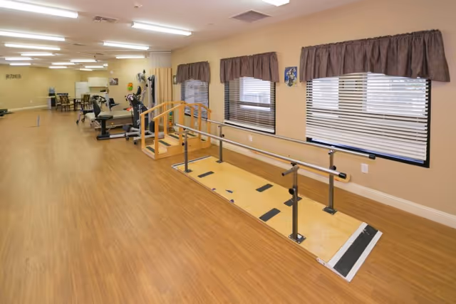 A spacious rehabilitation room with wooden flooring, featuring parallel bars and a small set of stairs for physical therapy exercises. The room has beige walls, three windows with brown curtains, and various exercise equipment in the background.