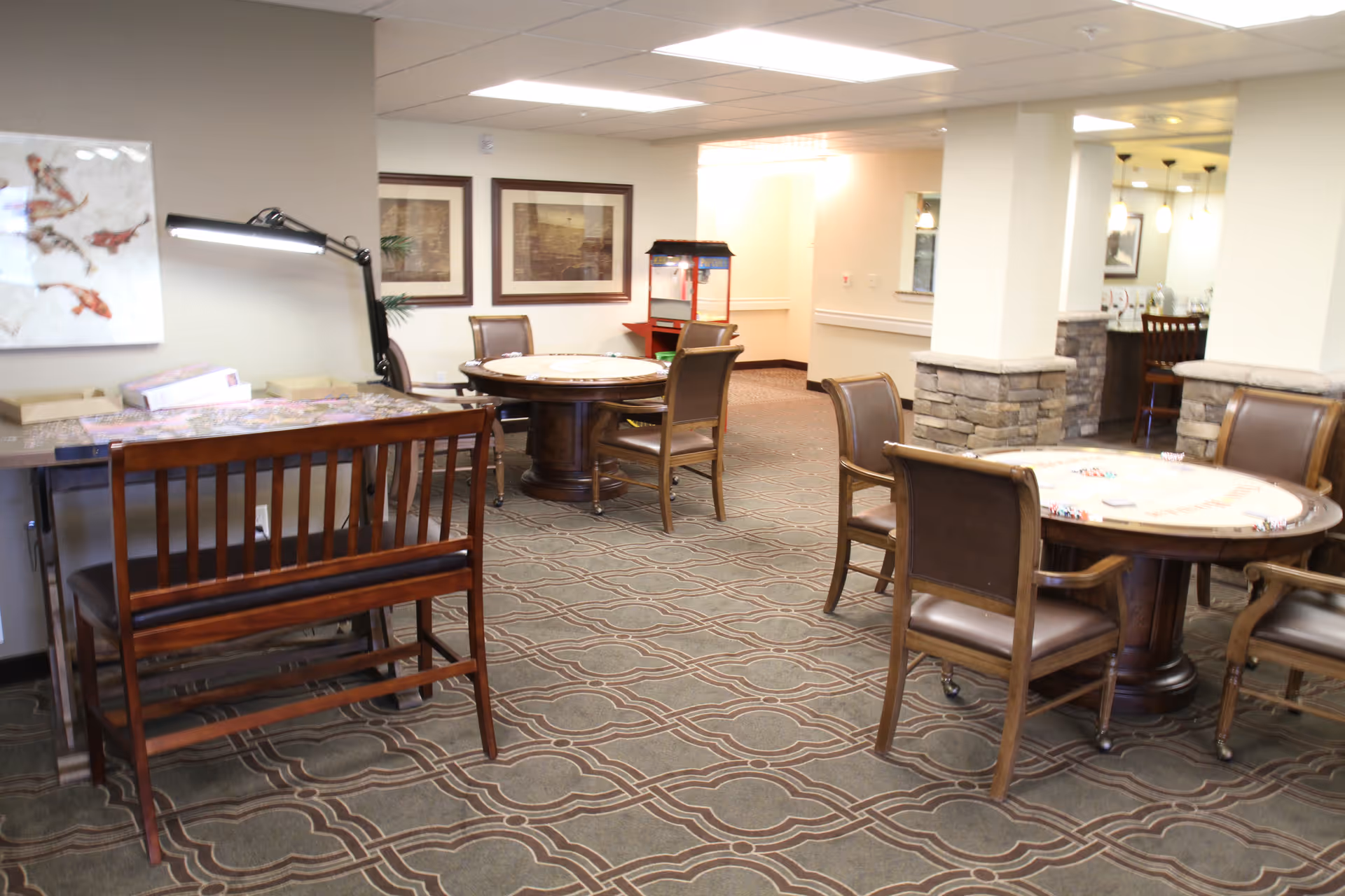Interior view of a common area in a senior living facility with round tables and chairs arranged for games or social activities. There is a wooden bench with a lamp on a side table, framed artwork on the wall, and a popcorn machine in the background near a kitchen or serving area.