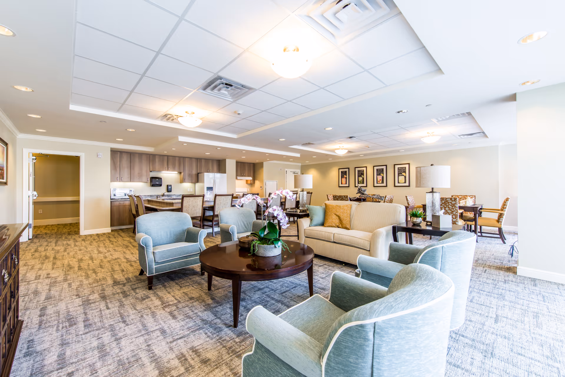 A spacious and well-lit common area in a senior living facility featuring a seating arrangement with a beige sofa, four light blue armchairs around a round wooden coffee table with a potted orchid, and additional dining tables and chairs in the background near a kitchen area with wooden cabinets and appliances.