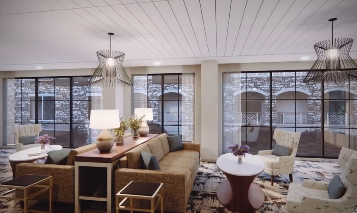 A bright communal lounge with sofas, armchairs, side tables, pendant lamps and large floor-to-ceiling windows overlooking a stone courtyard.