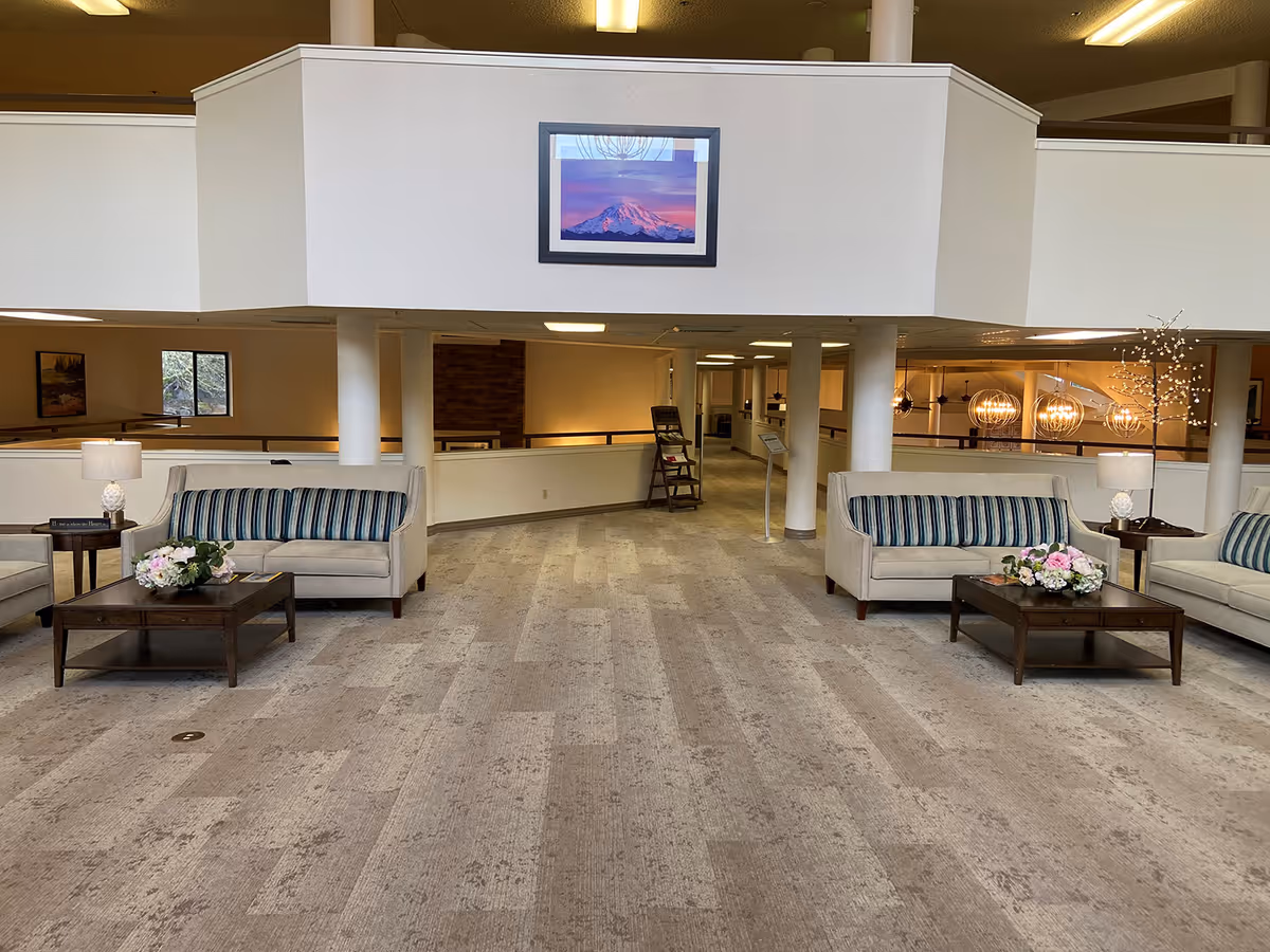 Spacious senior living facility common area with beige carpet flooring, two beige sofas with striped cushions, wooden coffee tables with flower arrangements, side tables with lamps, and a framed picture of a mountain on the wall above. The area is well-lit with ceiling lights and has a modern, clean design with white walls and pillars.