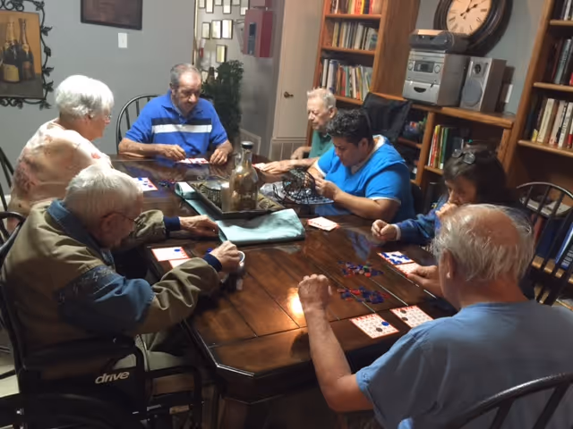 A group of elderly residents sitting around a table playing a bingo-style game in a cozy activity room.