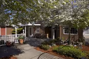 Outdoor view of a senior living facility courtyard with a concrete pathway, green shrubs, potted plants, and a tree with white blossoms. The building has brown siding and white trim around the windows and doors.