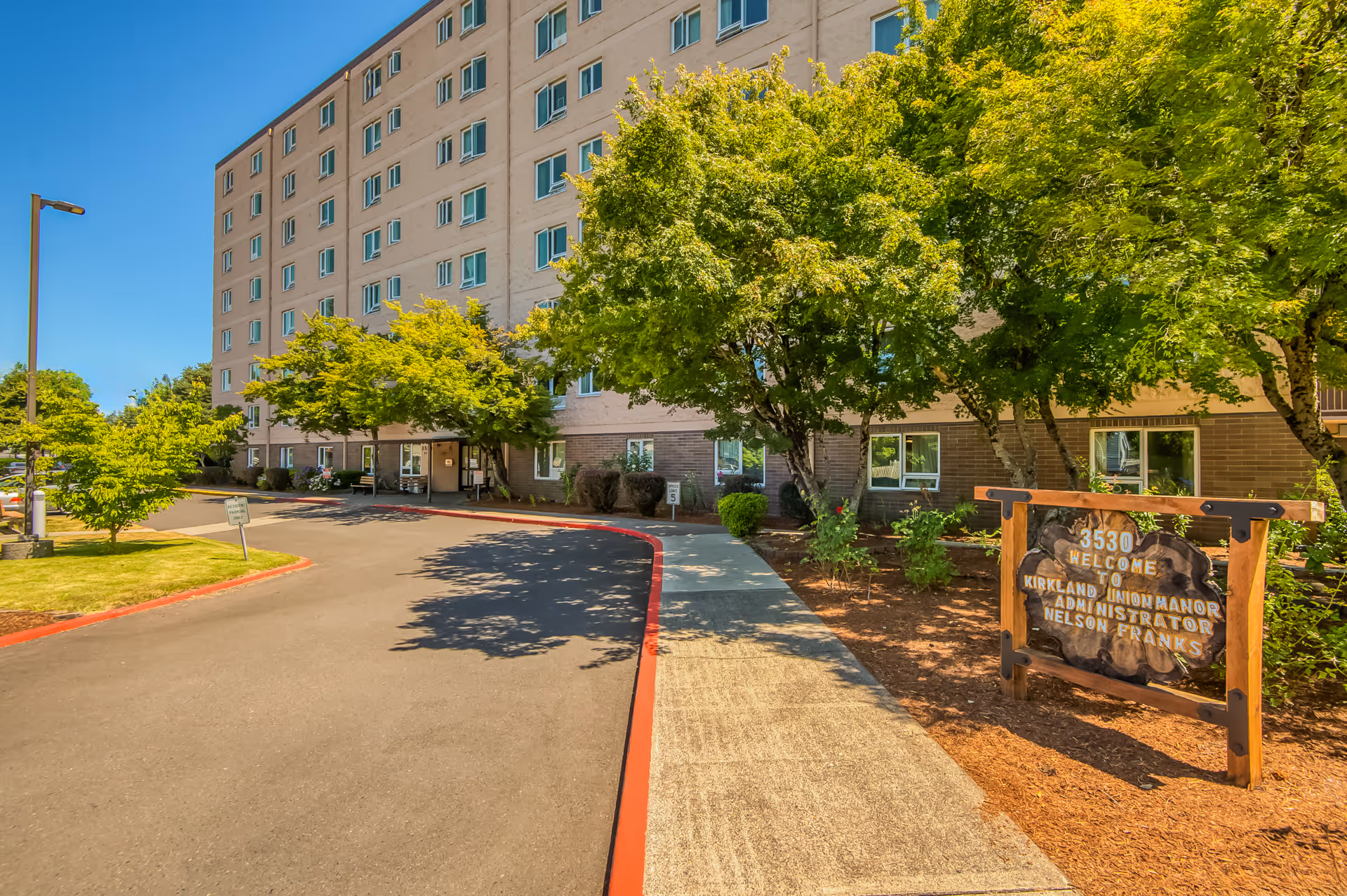 Front exterior of a multi-story senior living building with a driveway, trees, and a wooden welcome sign.