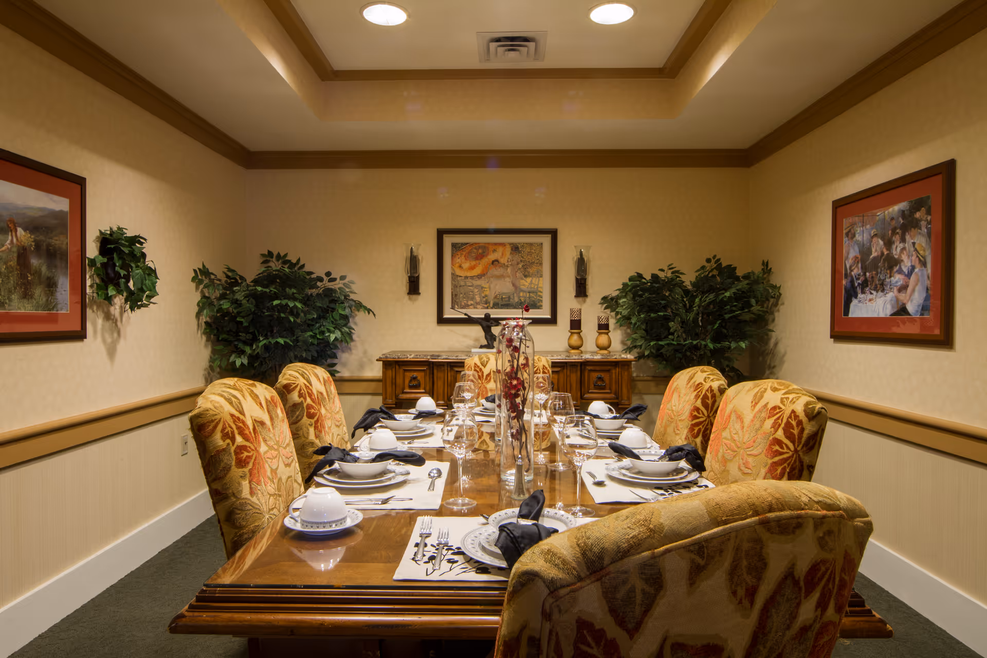 A formal dining room with a wooden table set for eight people. The table is arranged with plates, bowls, glasses, silverware, and black napkins. The chairs have a floral upholstery pattern. The room has beige walls with framed artwork, two large potted plants, and a wooden sideboard with decorative items. The ceiling has a recessed light fixture.