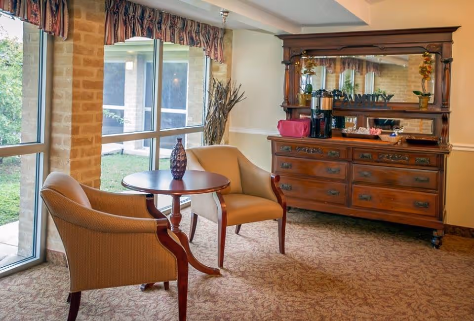 A cozy sitting area with two armchairs and a small round table by large windows and a wooden dresser with a mirror and coffee station.