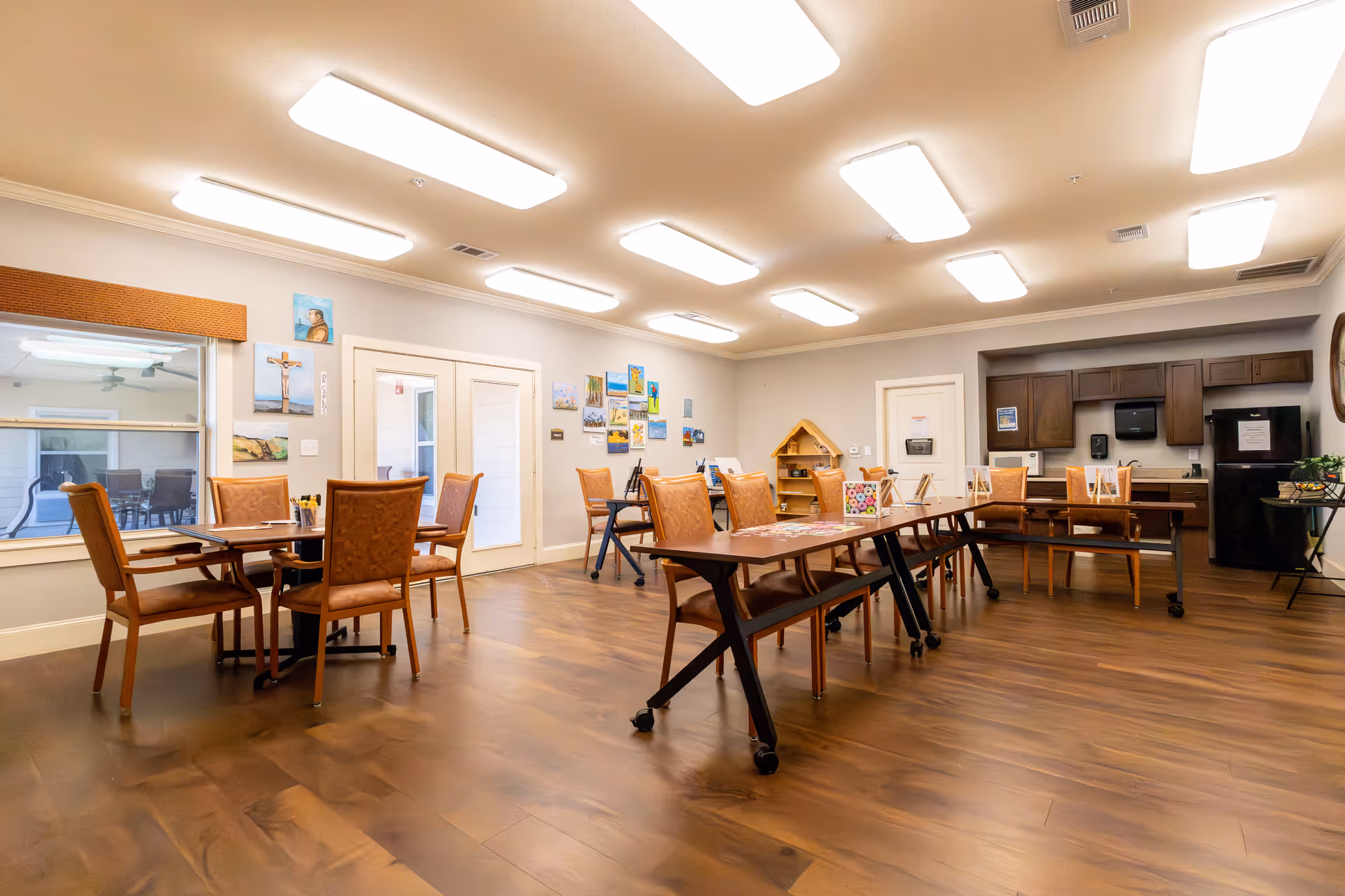 Spacious communal activity and dining room with tables and chairs, wall art, and a kitchenette area.