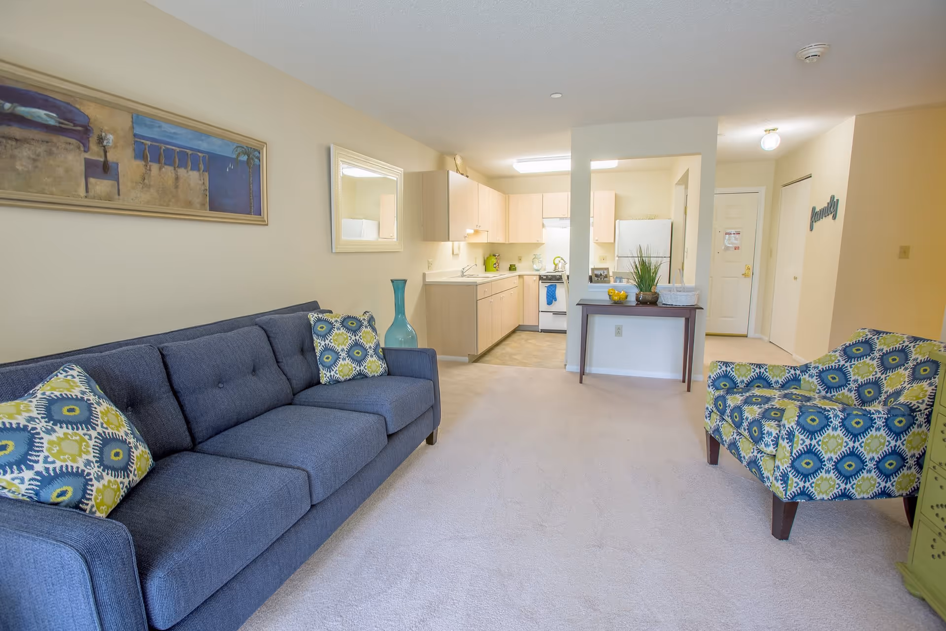 A bright and clean living room area with a blue sofa adorned with patterned cushions and a matching armchair. The room features light beige walls, a large framed painting, a mirror, and a small table with decorative items. In the background, there is a compact kitchen with light wood cabinets, a white refrigerator, stove, and sink. The entrance door and hallway are visible to the right.