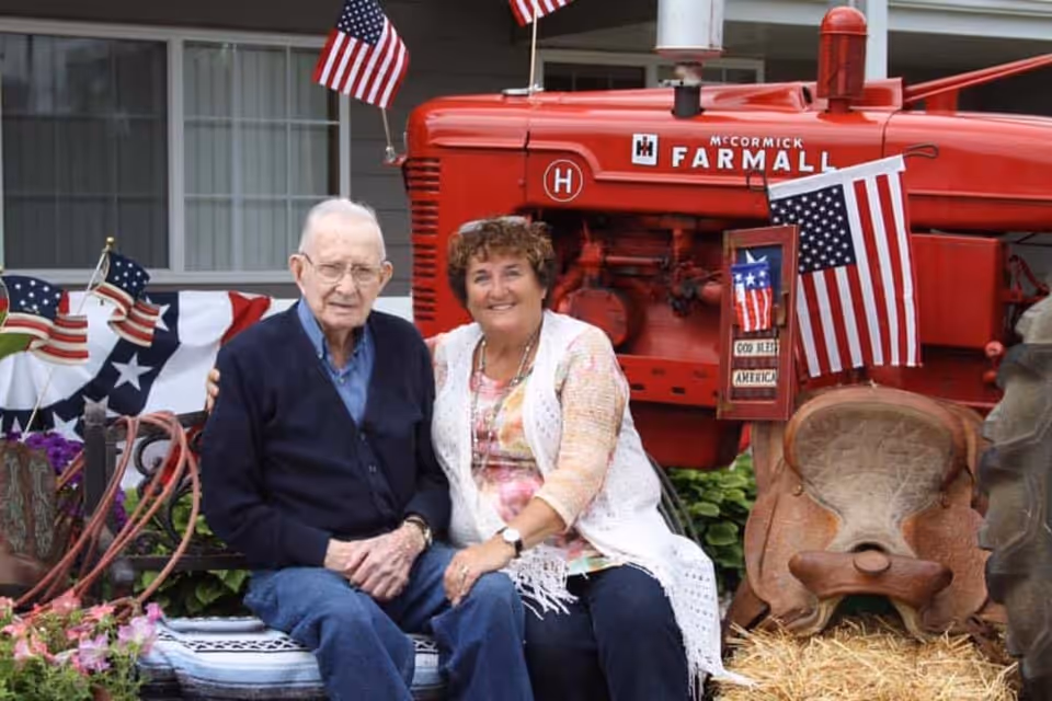 An elderly man and a middle-aged woman sitting together on a bench outdoors in front of a red McCormick Farmall tractor decorated with American flags and patriotic banners.