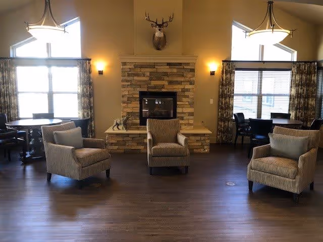 A cozy living room area with three beige armchairs arranged around a stone fireplace. Above the fireplace is a mounted deer head. There are two large windows with patterned curtains on either side of the fireplace, and round dining tables with chairs are visible near the windows. The room has warm lighting and wooden flooring.