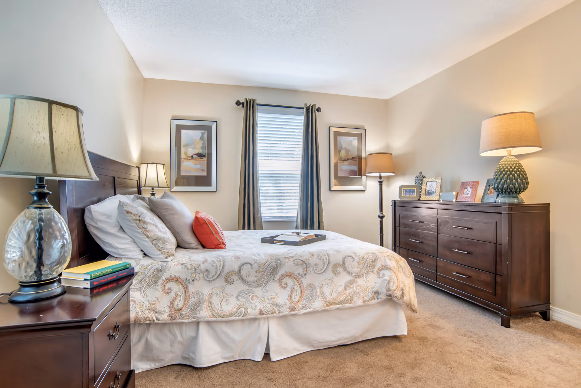 A bright bedroom with a bed featuring patterned bedding, nightstands and lamps, a dresser, and a window with curtains.