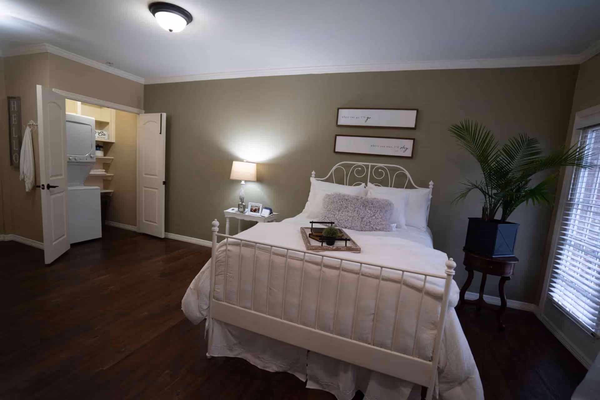 A furnished bedroom with a white metal bed, bedside table and lamp, potted plant, and an open closet showing a stacked washer-dryer.