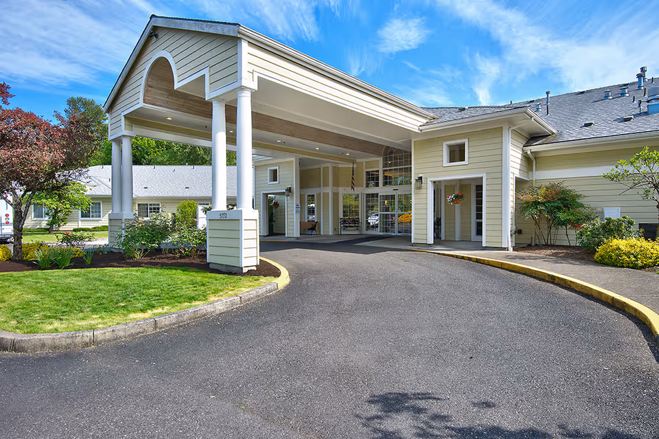 Front entrance of an assisted living building with a covered drive-through portico, white columns, landscaped grounds, and a curved driveway.