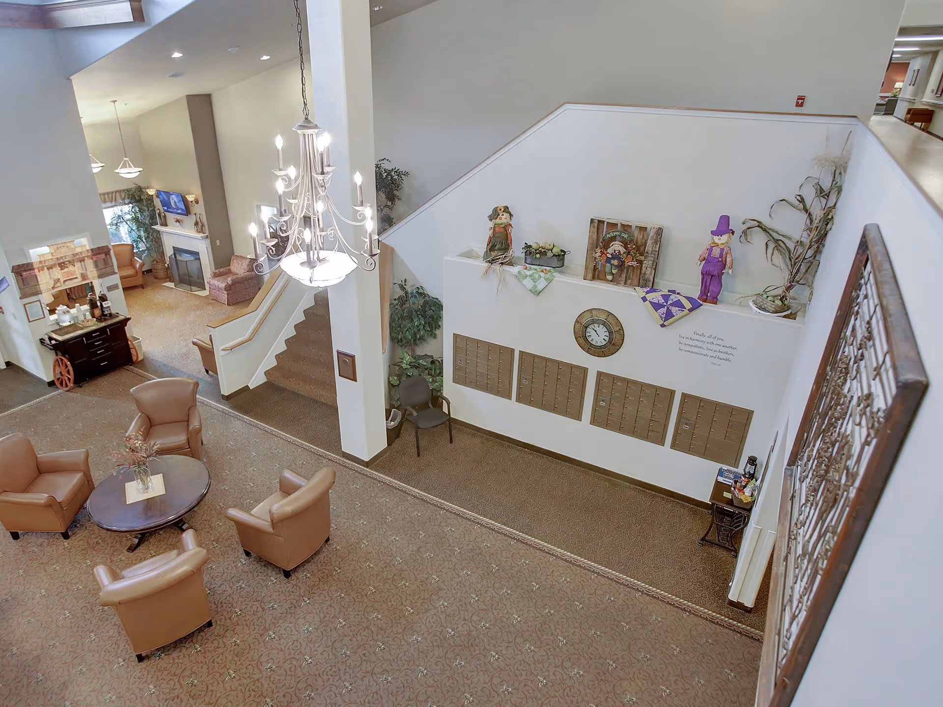 Interior view of a senior living facility common area with a seating arrangement of four tan leather chairs around a round wooden table. A chandelier hangs from the ceiling above. There is a staircase leading to an upper floor, a fireplace with a TV mounted above it, and decorative items on a ledge above mailboxes on the wall. The carpet is patterned and the walls are light-colored.