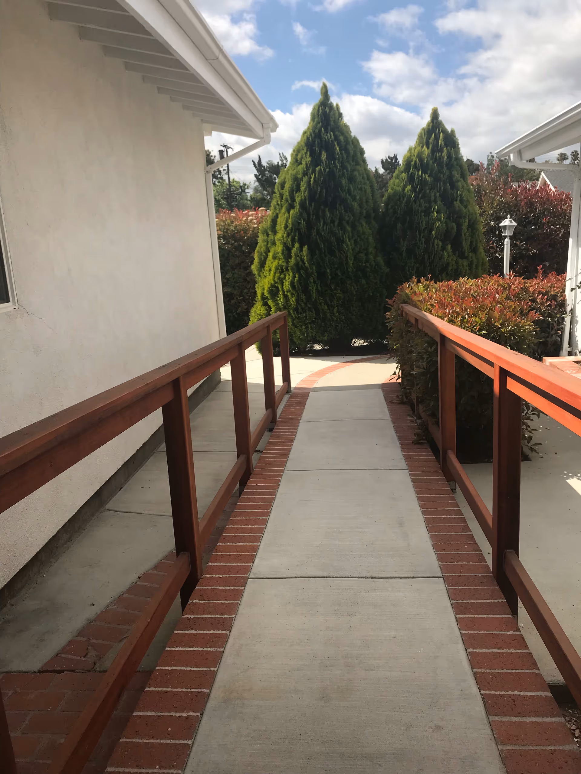 A concrete walkway with red brick edging and wooden handrails on both sides, leading between two white buildings towards green conical bushes and a partly cloudy sky.