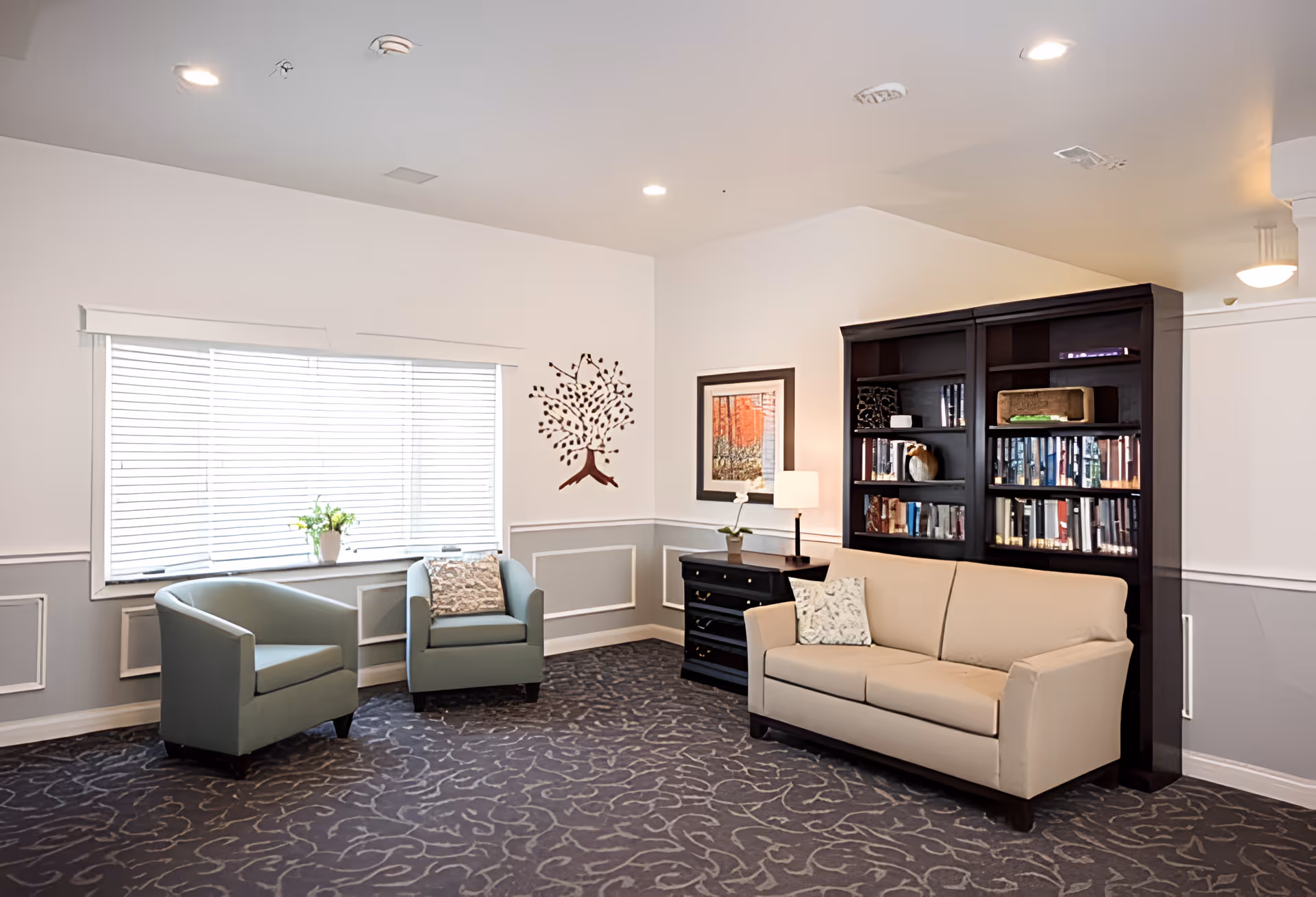 A cozy living room area with two light blue armchairs and a beige sofa. There is a large window with white blinds, a small black dresser with a lamp and a plant, a dark wooden bookshelf filled with books, and a framed picture on the wall. The walls are painted white with gray wainscoting, and the carpet has a subtle pattern.