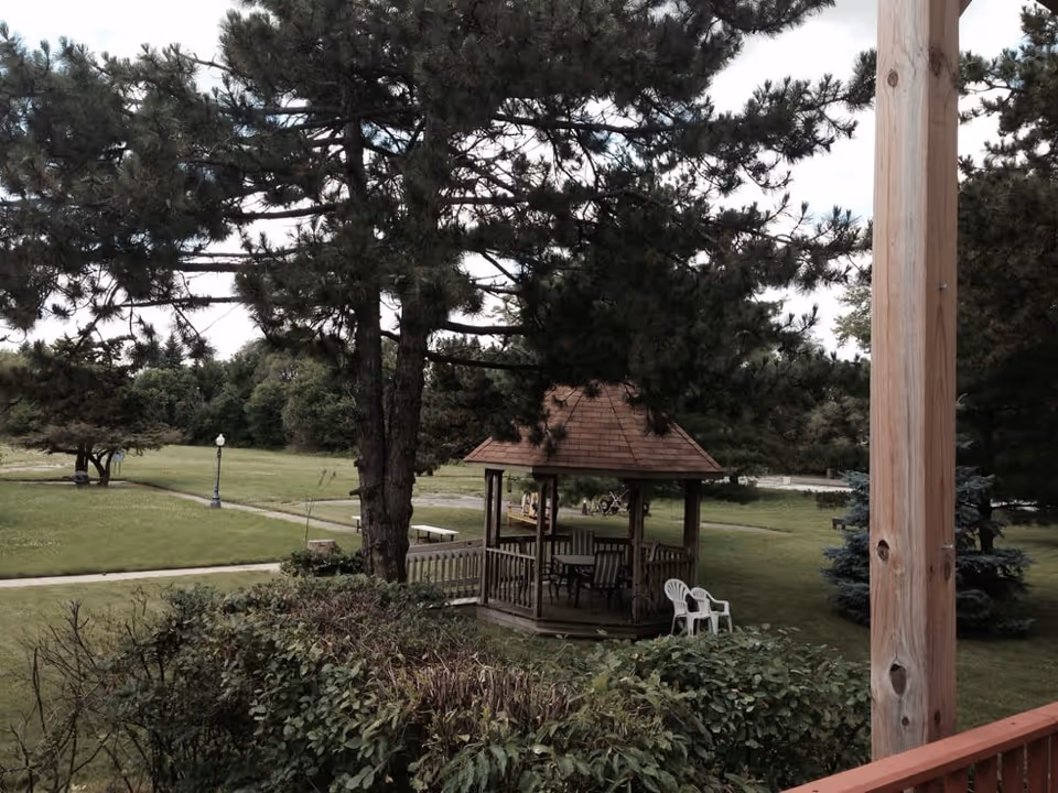 A peaceful outdoor area with a wooden gazebo surrounded by trees and greenery. There are several chairs inside the gazebo and two white plastic chairs outside it. A paved walkway and a lamppost are visible in the grassy area.