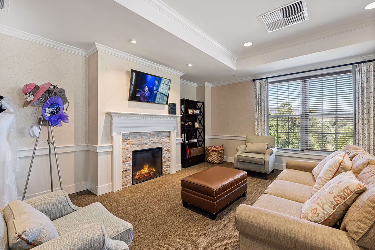 A cozy living room with a beige sofa adorned with patterned pillows, a matching armchair, and a brown leather ottoman in the center. A fireplace with a stone surround and white mantel is lit, above which a flat-screen TV is mounted. To the left, a coat rack holds several colorful hats and a white garment. A large window with blinds and patterned curtains lets in natural light, revealing greenery outside. A dark wooden bookshelf and a woven basket are placed in the corner.