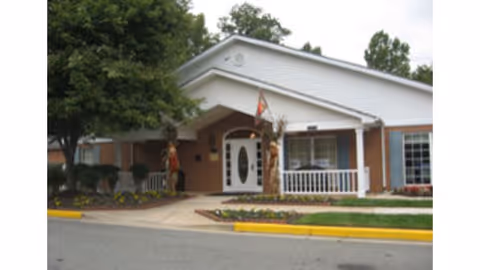Exterior view of a single-story brick building with white trim and a gabled roof. The entrance features a covered porch with white railings and a white door with oval glass panels. There are two carved wooden statues on either side of the entrance, a flag hanging above the door, and landscaping with trees and flower beds in front.