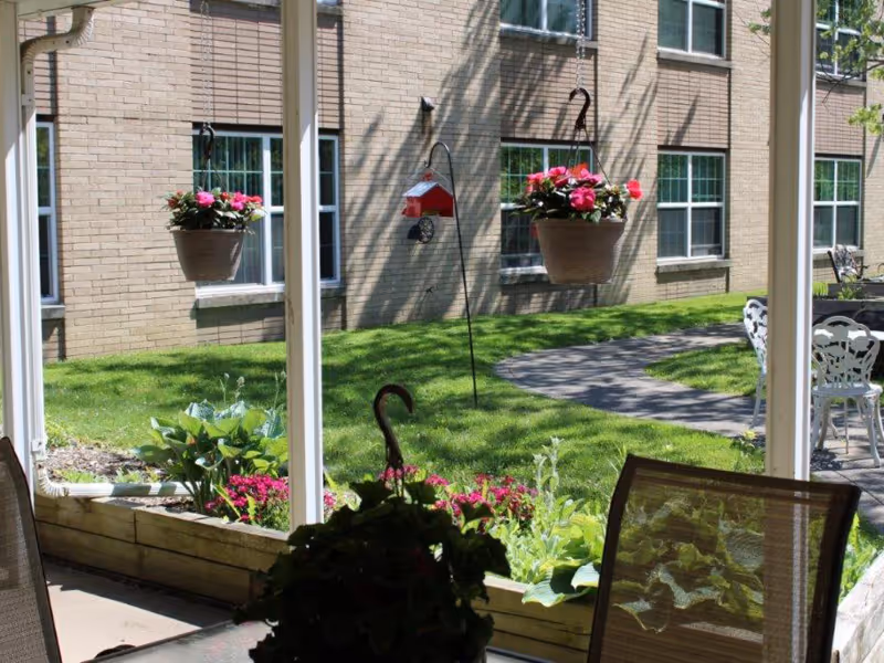 View from a covered patio area looking out onto a green lawn with hanging flower pots, a bird feeder, and a curved walking path. The background shows a brick building with multiple windows. There are outdoor chairs and a table visible on the right side.
