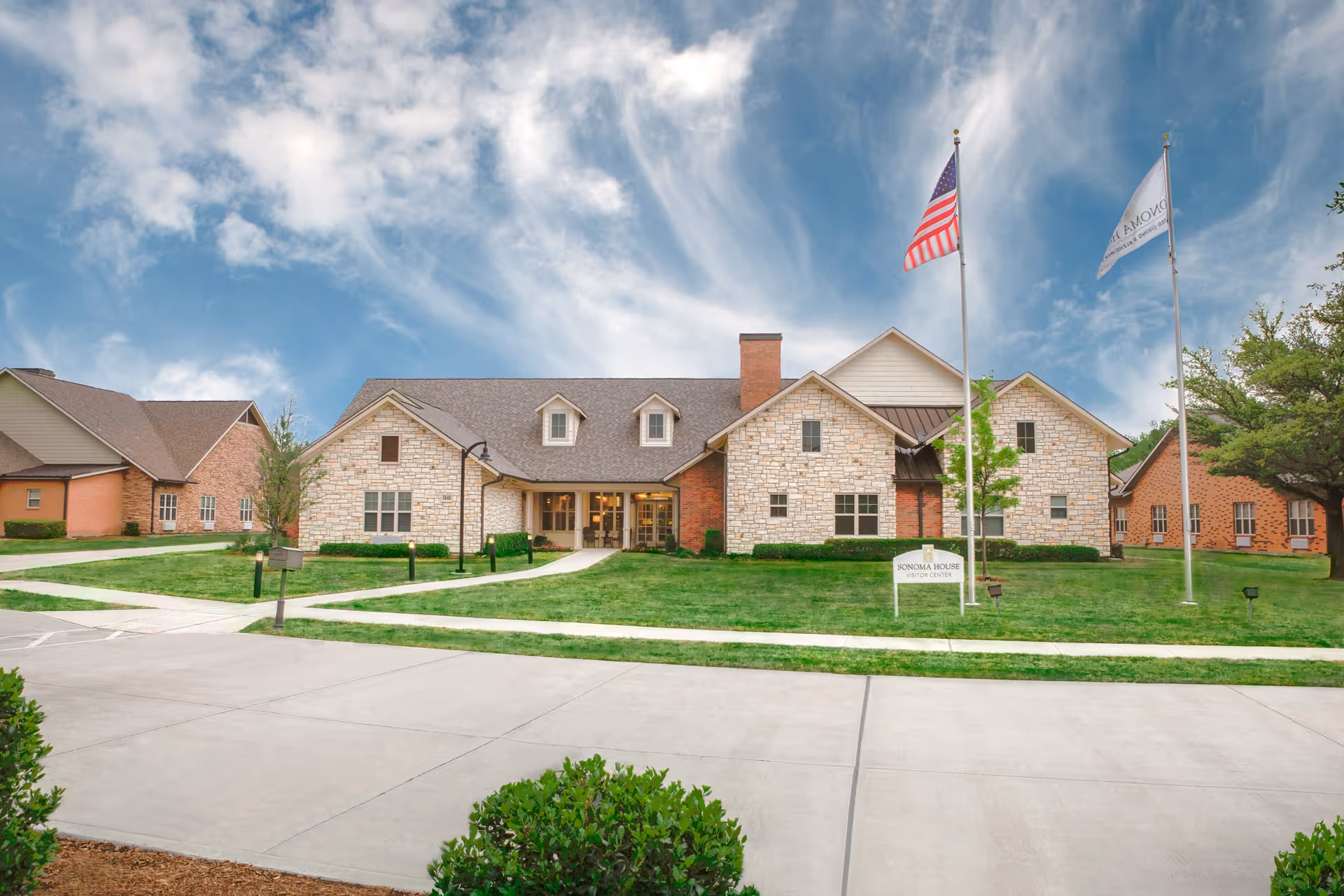 Front exterior of a stone-and-brick assisted living building with two flagpoles, a small sign, and a manicured lawn under a blue sky.