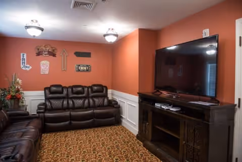 Cozy living room with leather recliner sofas, a large flat-screen TV on a wooden cabinet, and orange walls with decorative wall hangings.