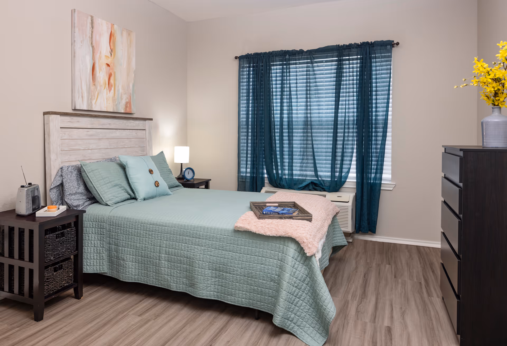 A bedroom with a bed covered in a light green quilt and matching pillows. There is a wooden headboard, a nightstand with a lamp and clock, a window with teal curtains, a dark wooden dresser with a vase of yellow flowers, and a tray with books on the bed. The floor is wood laminate.