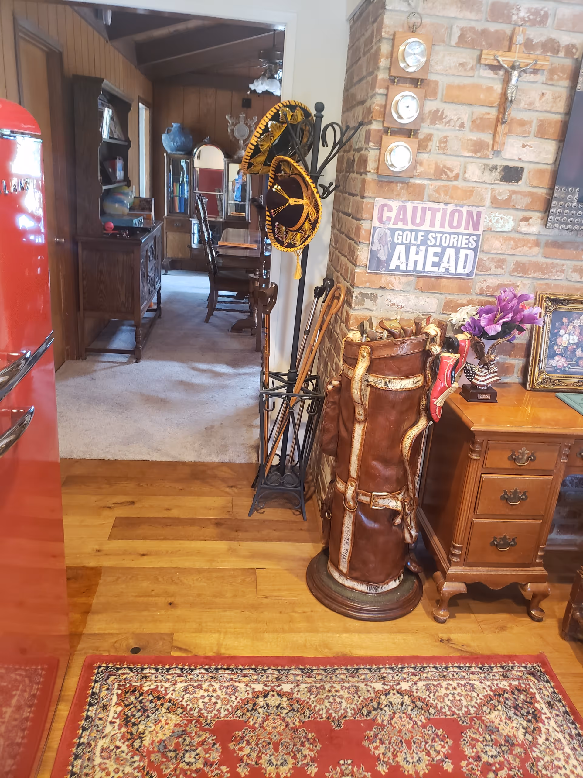 Interior view of a home entry/hallway showing a decorative golf bag, coat rack with sombreros, a brick wall with a sign reading "CAUTION GOLF STORIES AHEAD", wooden furniture and a rug leading into a dining room.