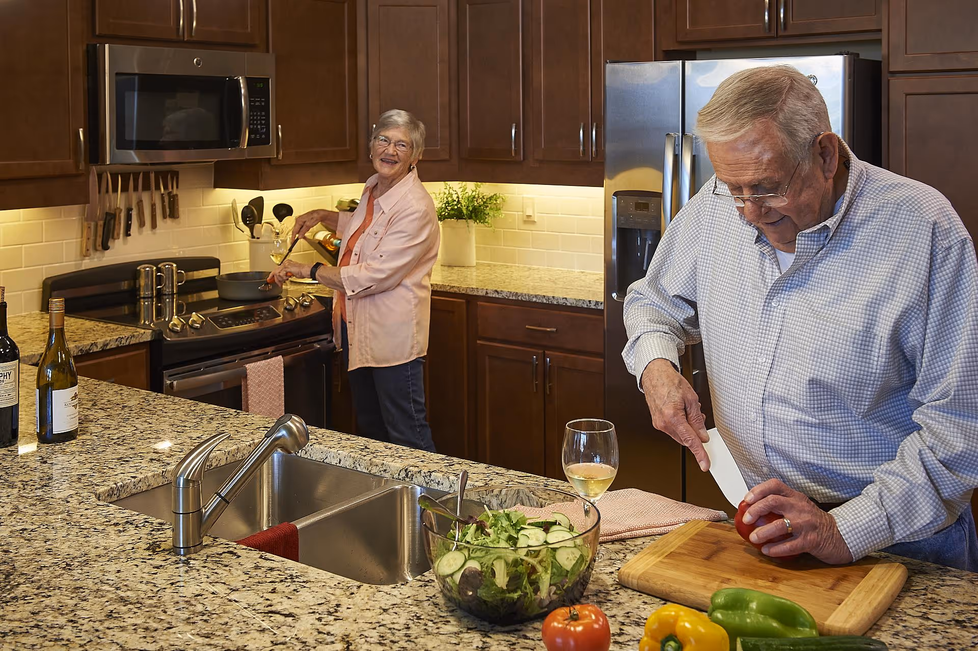 An elderly man and woman preparing food together in a modern kitchen. The man is slicing a tomato on a wooden cutting board, while the woman is cooking on the stove. The kitchen features granite countertops, wooden cabinets, a stainless steel refrigerator, and a microwave. There is a glass bowl with salad and a glass of white wine on the counter.