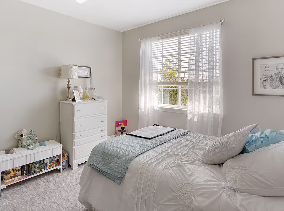 A bright bedroom with a neatly made bed featuring white bedding and a light blue throw blanket. There is a window with sheer white curtains letting in natural light. A white dresser with a lamp, framed pictures, and decorative items sits against one wall. A small white storage bench with board games and a stuffed animal is also visible. A framed drawing hangs on the wall beside the window.
