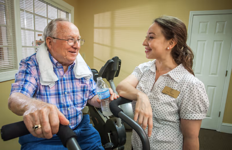 An elderly man with a towel around his neck is sitting on an exercise bike holding a water bottle, smiling and talking with a female staff member wearing a name tag in a well-lit room with beige walls and a white door.