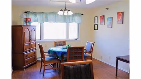 A dining area with a round table covered with a blue tablecloth and four wooden chairs around it. There is a wooden cabinet on the left side and a window with a blue valance curtain letting in natural light. The walls are decorated with framed artwork, and the floor is wooden.
