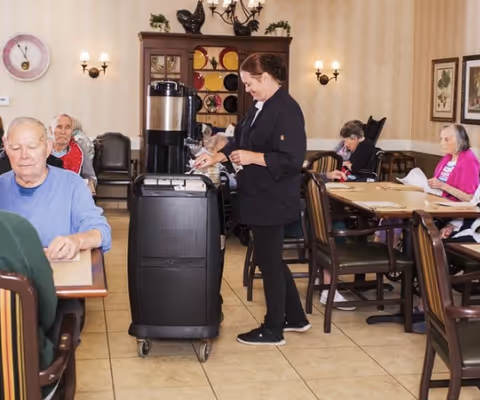 A staff member serving beverages from a cart while seniors sit at tables in a dining room.