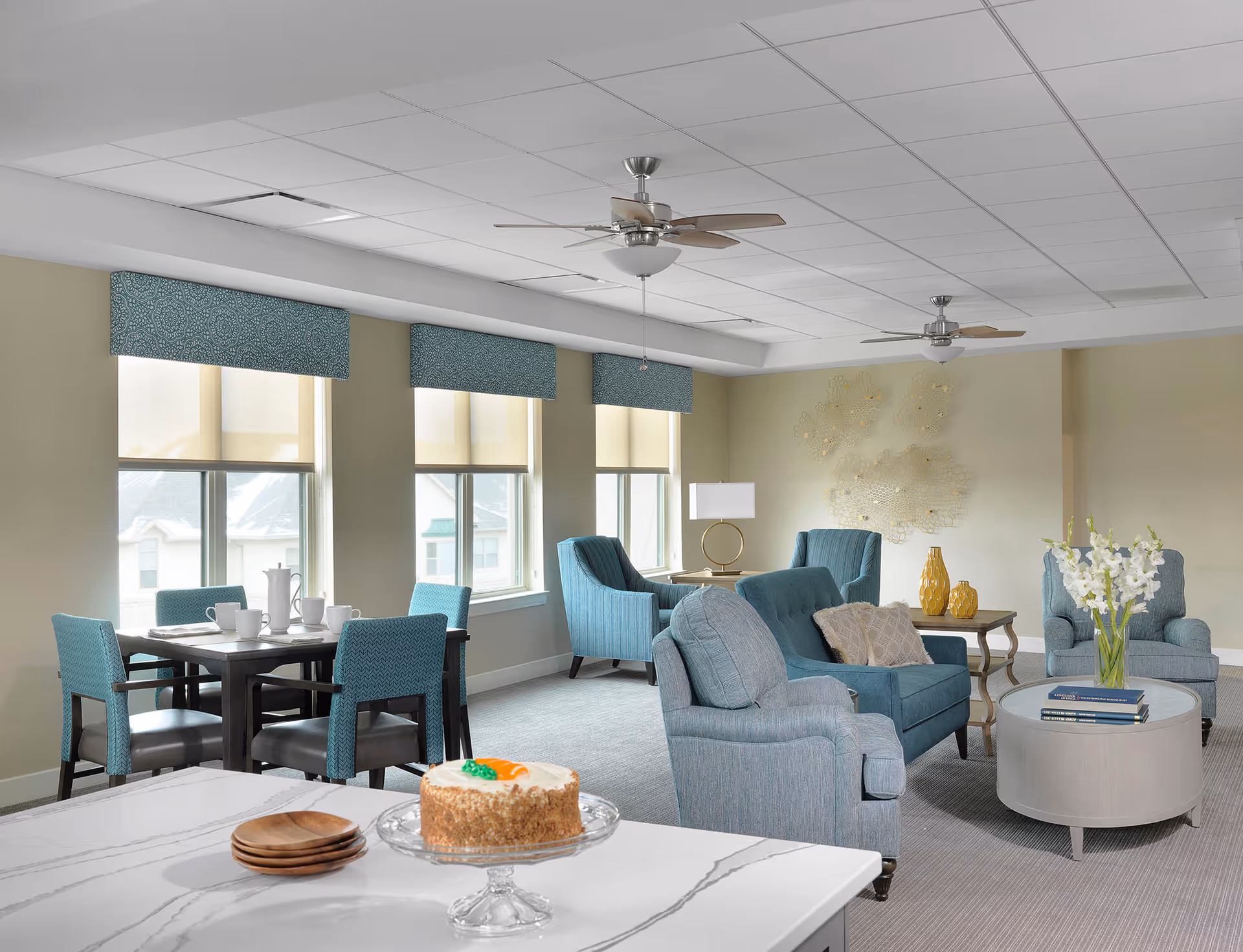 Bright communal lounge with teal upholstered chairs, a dining table by windows, and a cake on a counter in the foreground.