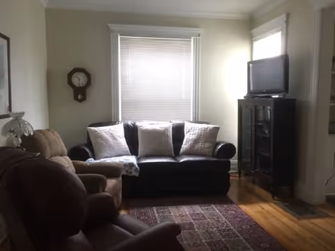 A cozy living room with a black leather sofa adorned with three white pillows, two brown recliner chairs, a patterned area rug on a wooden floor, a wall clock, and a TV on a dark wooden cabinet near a window with blinds.