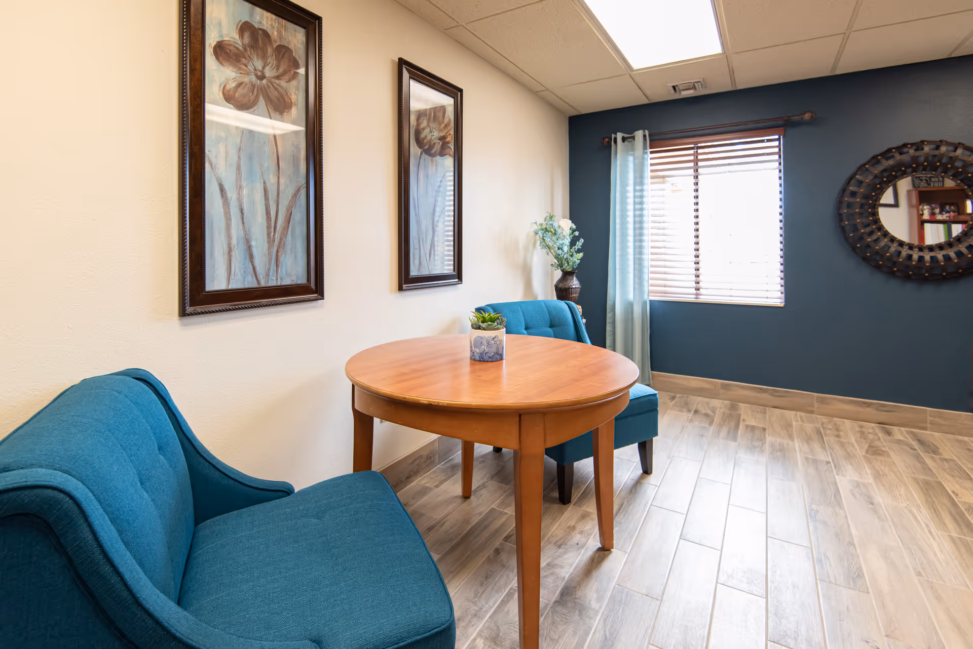A cozy sitting area with a round wooden table and two teal upholstered chairs. The room has light wood flooring, a window with wooden blinds and light blue curtains, two framed floral paintings on a beige wall, and a decorative round mirror on a dark blue accent wall.