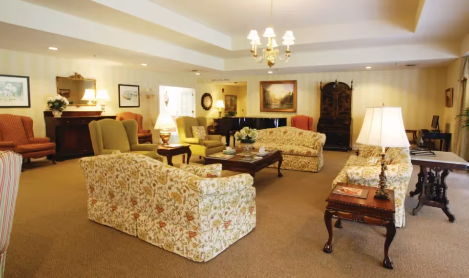 A spacious living room with floral patterned sofas, armchairs, wooden coffee tables, and lamps. The room features warm lighting, framed artwork on the walls, a piano in the background, and a carpeted floor.
