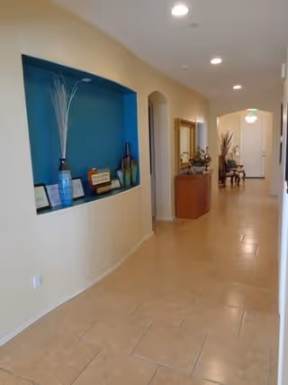 A well-lit hallway with beige tiled flooring and light yellow walls. On the left side, there is a recessed blue niche displaying decorative items including vases and framed pictures. Further down the hallway, there is a wooden console table with a mirror above it and some decorative plants. The hallway leads to a white door at the end.