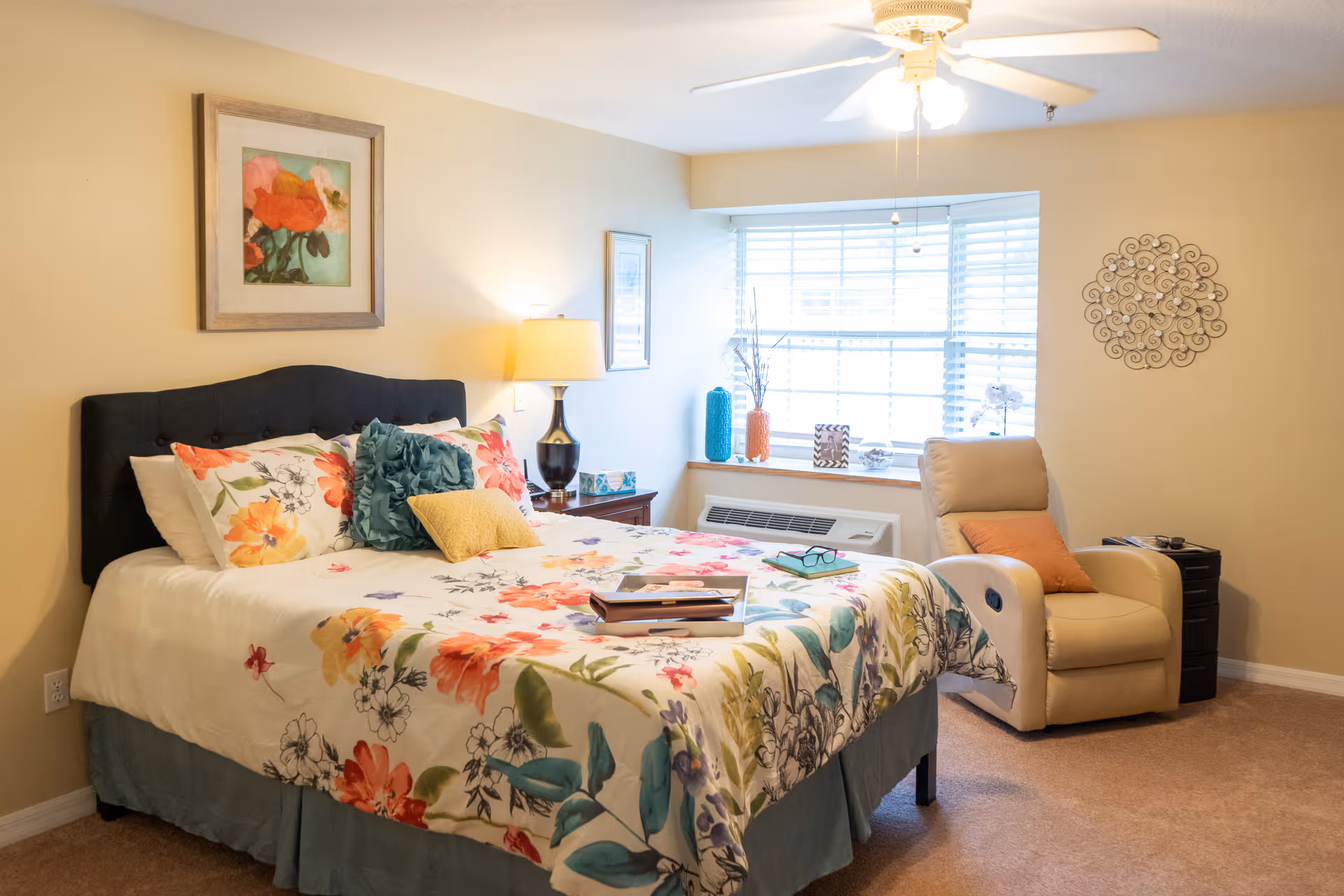 A cozy bedroom with a bed featuring a floral bedspread and multiple pillows. There is a nightstand with a lamp and decorative items next to the bed. A large window with blinds lets in natural light. A comfortable beige recliner chair with an orange pillow is positioned near the window. The walls are decorated with framed artwork and a metal wall decoration. The room has beige carpeting and a ceiling fan with lights.