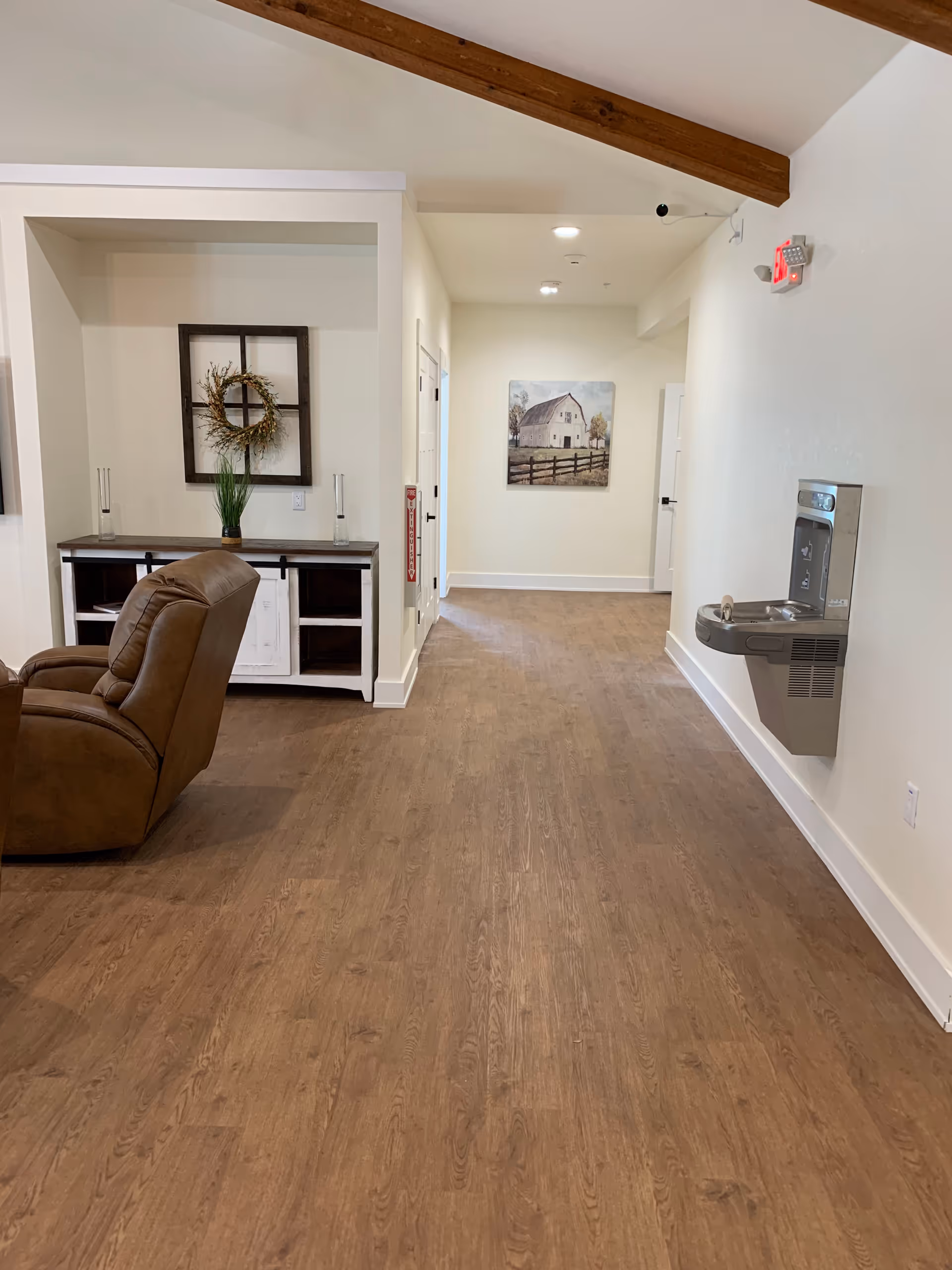 Interior hallway of an assisted living facility with wooden flooring, a brown recliner chair on the left, a console table with decorative items including a wreath and a plant, a water fountain mounted on the right wall, and a painting of a barn on the far wall under a ceiling with exposed wooden beams.