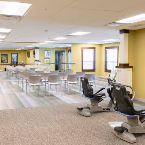 A spacious senior living facility room with exercise equipment in the foreground and rows of chairs arranged in the background. The room has yellow walls, large windows with blinds, and framed artwork on the walls. The floor has a mix of carpet and patterned vinyl tiles.