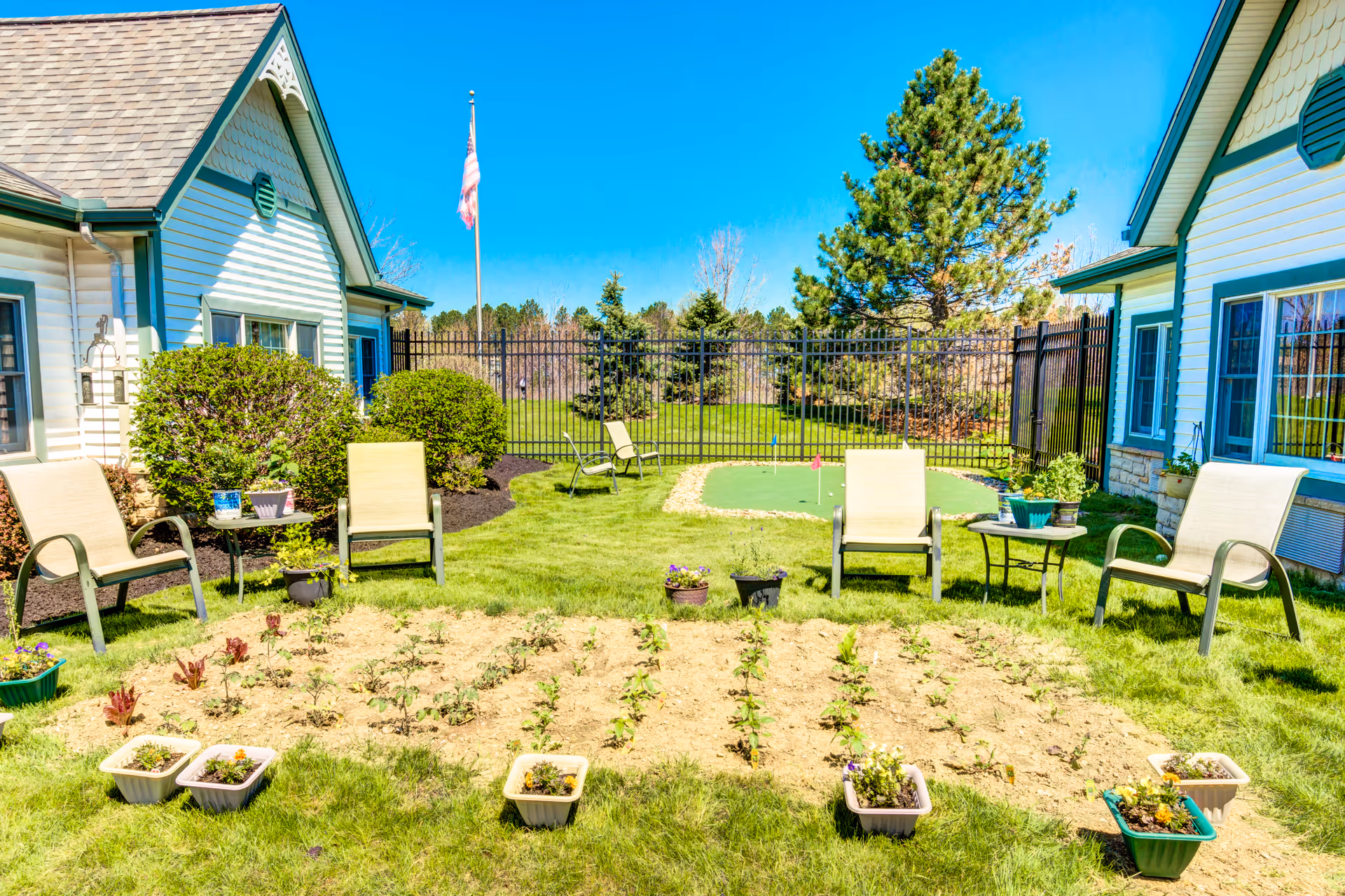 A sunny outdoor garden area at Symphony at Mentor featuring a small vegetable or flower garden with several potted plants around it. There are four beige patio chairs and two small tables with potted plants on them. The garden is surrounded by two white buildings with green trim, a black metal fence, and an American flag on a flagpole. Trees and a clear blue sky are visible in the background.
