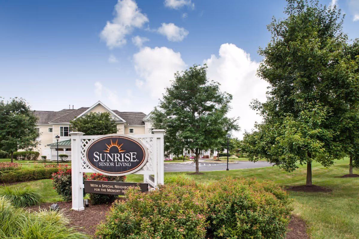 Exterior view of Sunrise Senior Living facility with a large sign in the foreground surrounded by bushes and trees, a parking lot, and a building with white siding and a gray roof in the background under a partly cloudy sky.
